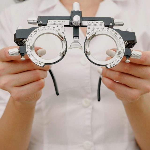 Eine Person in einem weißen Laborkittel, wahrscheinlich ein Augenarzt, hält ein Paar Testgläser, die für Sehdiagnostik oder Kataraktuntersuchungen verwendet werden, mit beiden Händen vor der Brust. Das Gesicht der Person ist nicht zu sehen.